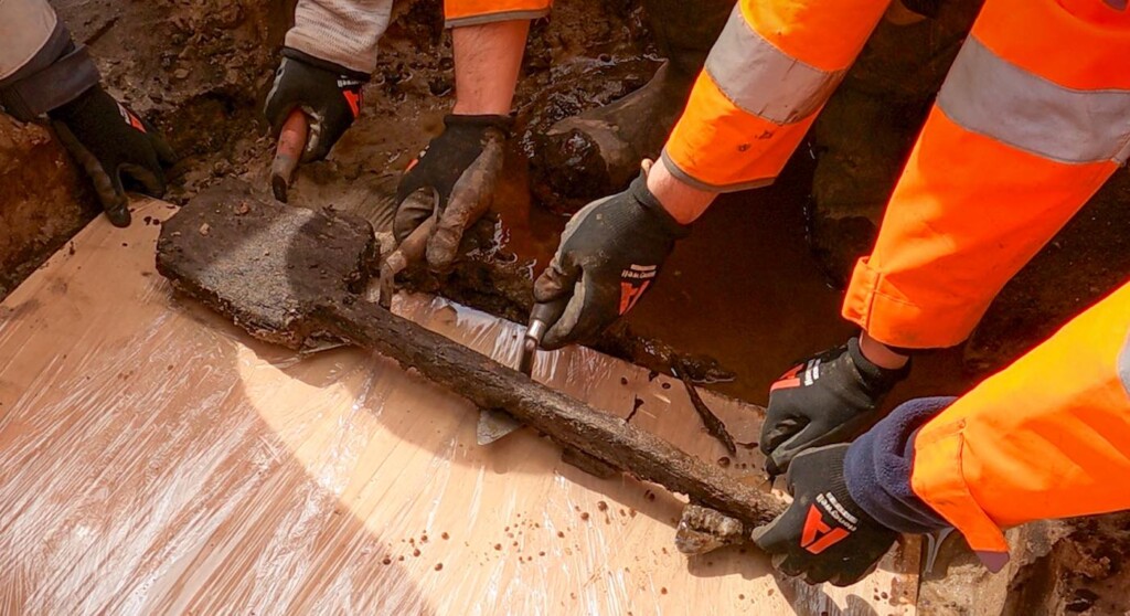 Archaeologists Carefully Lift the Fragile Spade From the Ground Onto a Board–Wessex Archaeology For the Moors at Arne Project at Poole Harbour Via Swns